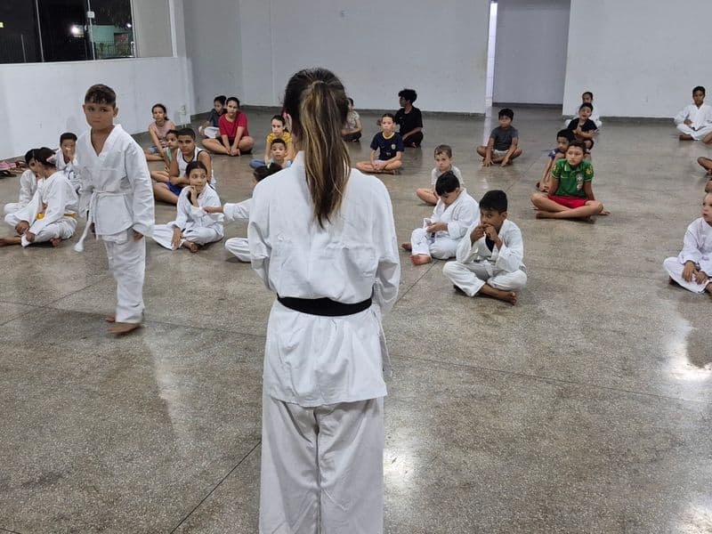 Crianças e adolescentes do projeto Realização participam de uma oficina de karatê em um amplo salão. Uma instrutora de costas, vestindo kimono branco e faixa preta, orienta o grupo. Alguns alunos estão em pé vestindo kimono, enquanto a maioria está sentada no chão prestando atenção.
