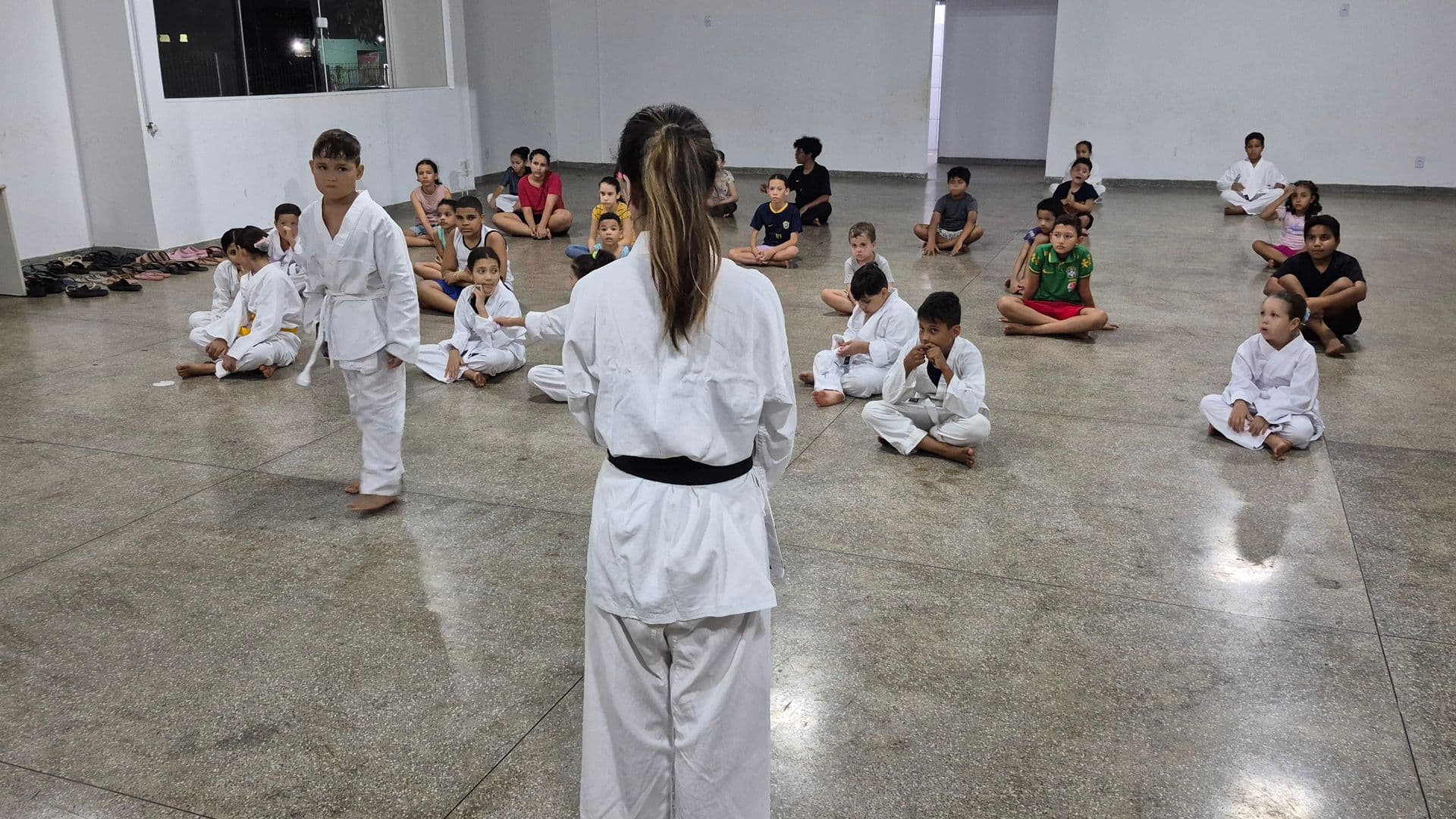 Crianças e adolescentes do projeto Realização participam de uma oficina de karatê em um amplo salão. Uma instrutora de costas, vestindo kimono branco e faixa preta, orienta o grupo. Alguns alunos estão em pé vestindo kimono, enquanto a maioria está sentada no chão prestando atenção.