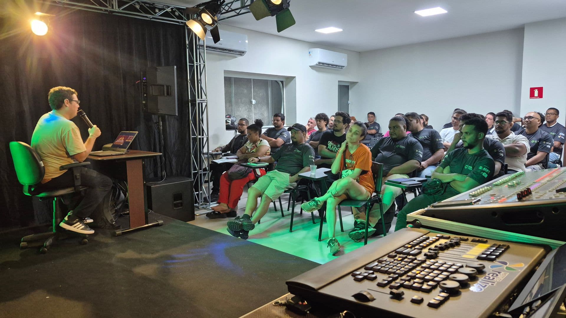 Imagem da Sala de aula com o professor falando ao microfone e os alunos sentados em carteiras ouvem atentamente.