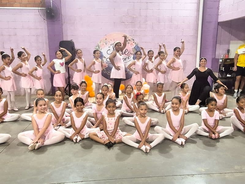 Grupo de meninas alunas da oficina de ballet em uma pose para foto vestidas com collants e sapatilhas, a frente meninas sentadas em pose de borboleta e atrás em pé meninas em pose de ballet, a direita do grupo a instrutora vestindo roupas pretas.