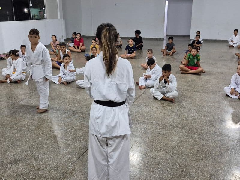 Crianças e adolescentes do projeto Realização participam de uma oficina de karatê em um amplo salão. Uma instrutora de costas, vestindo kimono branco e faixa preta, orienta o grupo. Alguns alunos estão em pé vestindo kimono, enquanto a maioria está sentada no chão prestando atenção.