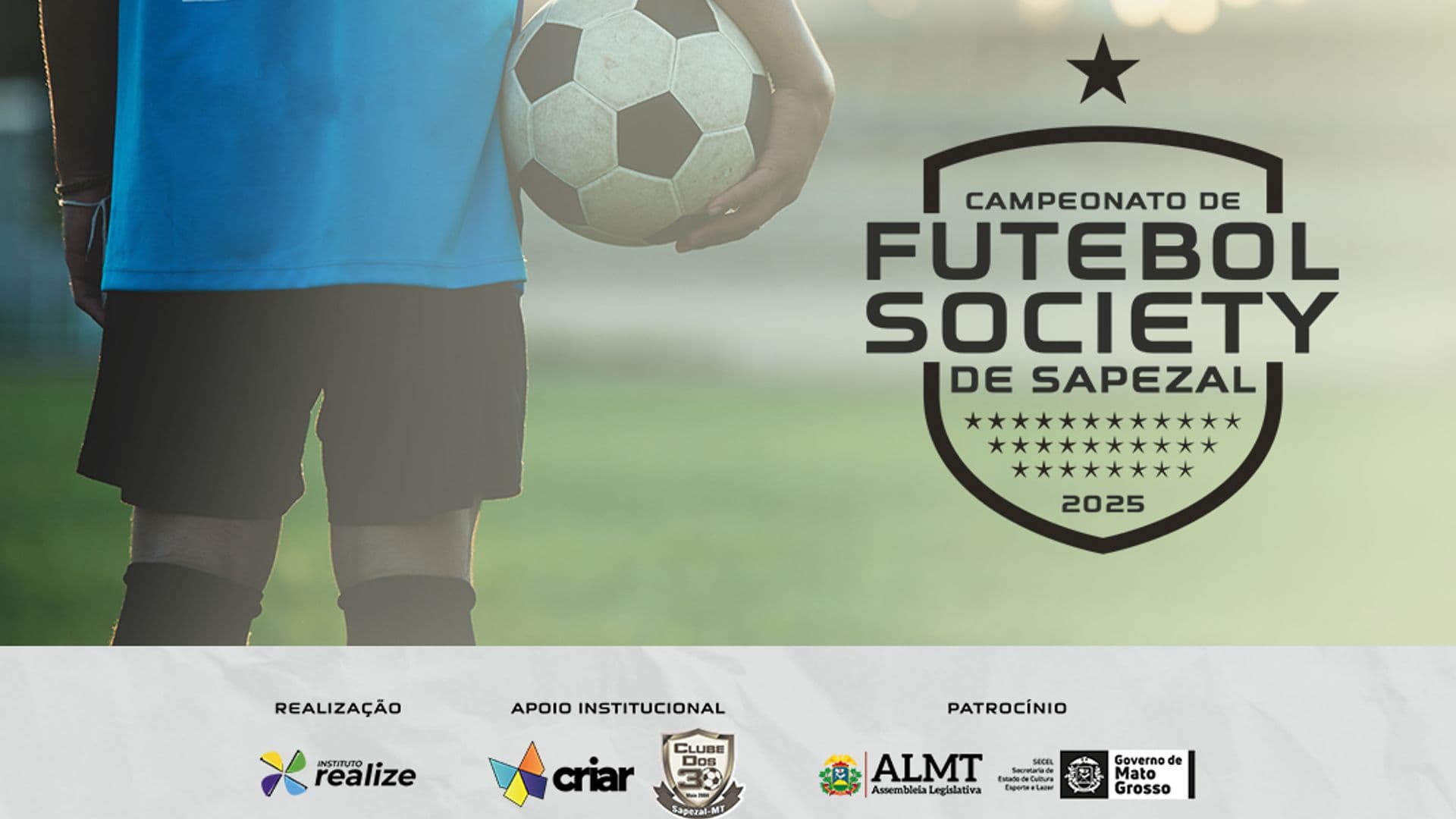 Foto de um rapaz de costas com uniforme de um time, segurando uma bola de futebol em pé dentro de um campo gramado com desfoque da imagem.